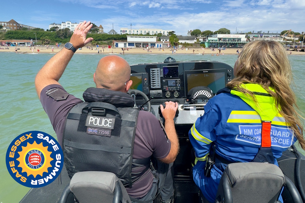 A uniformed Essex Police marine officer and a member of HM Coastguard's Search and Rescue team in a police patrol boat, waving towards a busy beach