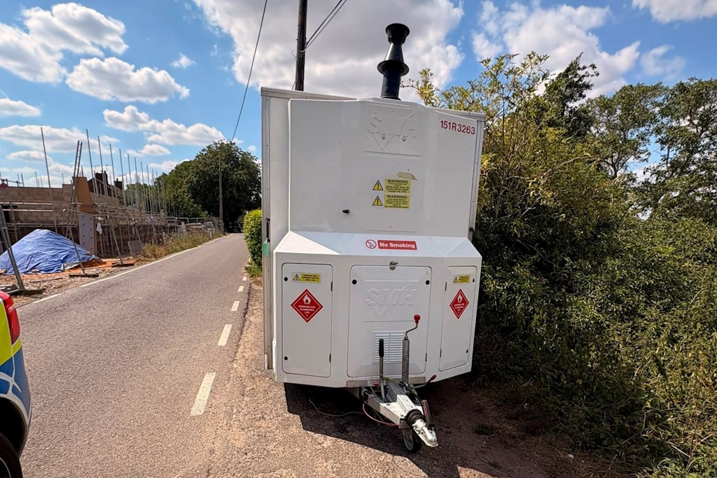 A white asbestos decontamination unit with hazard warning signs parked on the side of a rural road next to a hedge