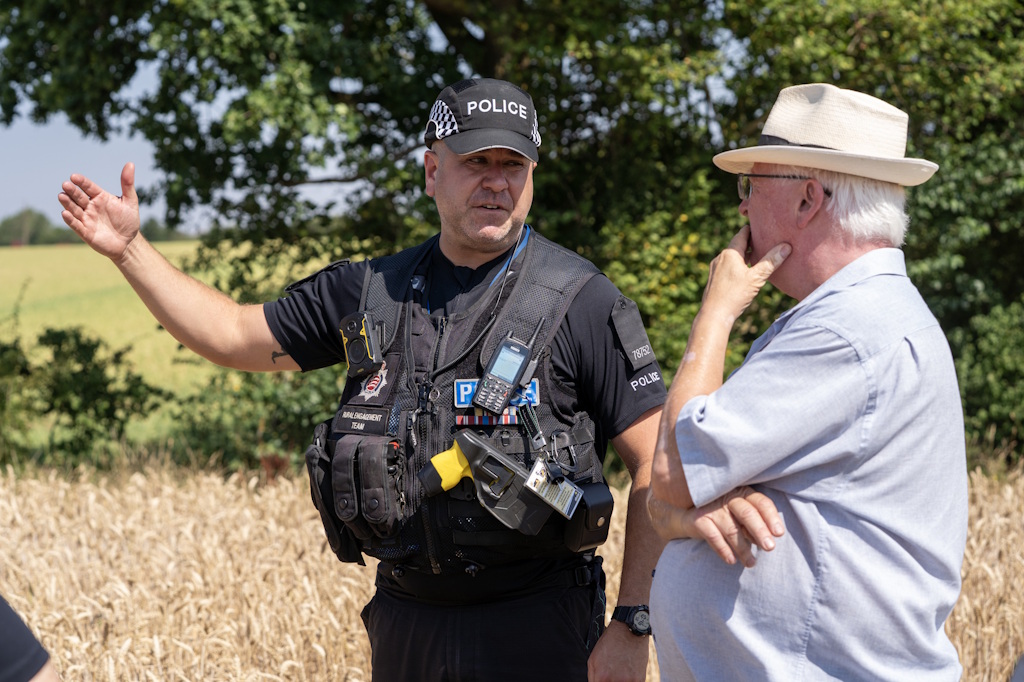 A uniformed Essex Police rural engagement officer speaks to a man in a straw hat standing at the edge of a wheat field