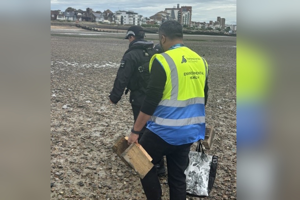 A uniformed Essex Police marine officer and a Southend City Council environmental health officer walk up a muddy beach, carrying wooden boxes and shopping bags