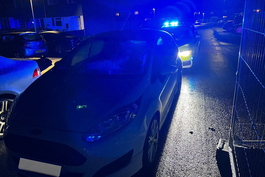 A night scene with a white car parked in front of an Essex Police liveried vehicle with flashing blue emergency lights