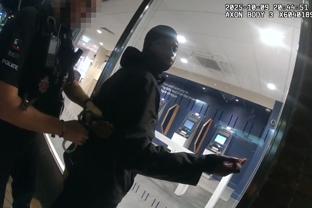 Essex Police body-worn footage still of a uniformed Essex Police neighbourhood policing officer preparing to handcuff a man in front of an ATM foyer, at night