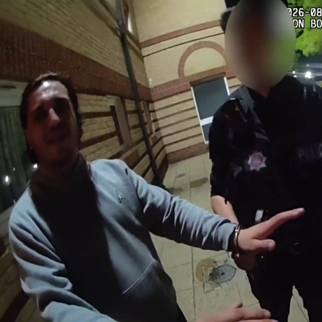 A man in a grey jumper with a handcuff on his wrist stood next to a uniformed police officer