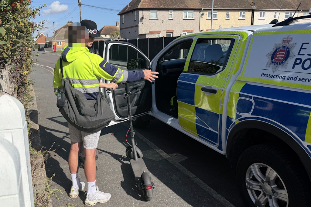 A uniformed Essex Police neighbourhood policing officer speaks with a person next to an e-scooter next to a marked Essex Police vehicle on a street