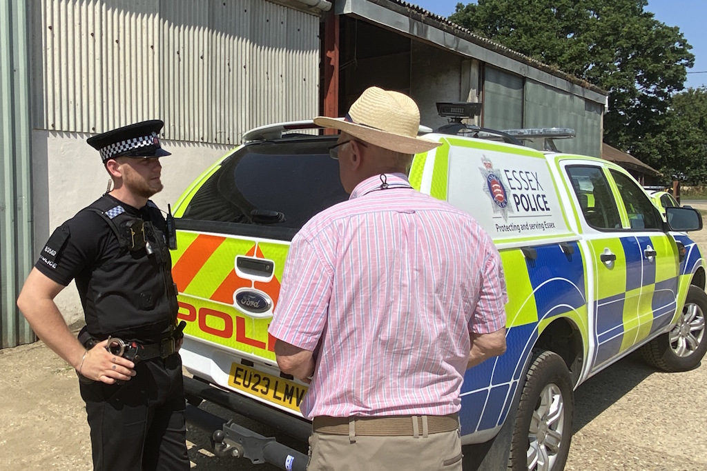 A uniformed Essex Police neighbourhood policing officer speaks to a man in a pink stripey shirt and a straw hat in a farmyard. They are standing in front of an Essex Police liveried vehicle, which is parked in front of a corrugated iron barn